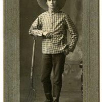 Portrait of a young man. The man wears a wide brim straw hat and a checked shirt. He is leaning against a pitchfork.