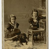 Portrait of two young girls and a small dog. The girls are both dressed in sailor suit style dresses.