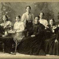 Portrait of a man and six women seated on wicker chairs.