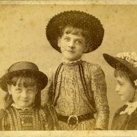 Portrait of three young girls. The girls all wear hats and patterned dresses.