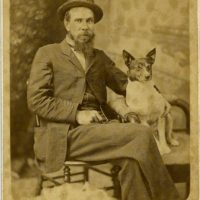 Portrait of a man sitting beside a small dog. The man has a beard and is dressed in a suit. The man and the dog are seated side by side and the man has his arm around the dog.