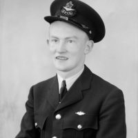 Black and white mid shot portrait of a man in an RAF or RCAF uniform. He is wearing a EFTS Instructor cap badge on his hat. There is also a small pin on his jacket. He is smiling. This image is on the left-hand side of a sheet of film containing two separate exposures.