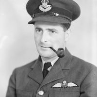 Black and white mid shot portrait of a man in an RAF uniform. There is an RAF Pilot's badge on his jacket and he is wearing a hat with an Officer's cap badge. He is posing with a pipe in the corner of his mouth.