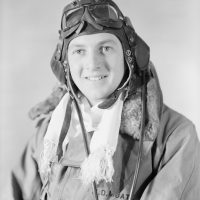 Black and white medium close up portrait of a man in a flight suit. He is wearing a coat with a fur collar and "D. Moate" written on it. He is also wearing a white scarf and a pilot's leather cap with goggles.