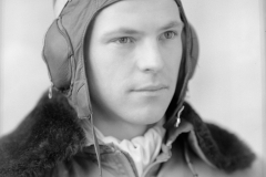 Black and white medium close up portrait of a man dressed as a pilot. His jacket has a fur collar and he is wearing a leather cap with goggles on top of his head. A white scarf is visible underneath his zipped up jacket. The negative (labelled a) was accompanied by one black and white photographic print (labelled b).